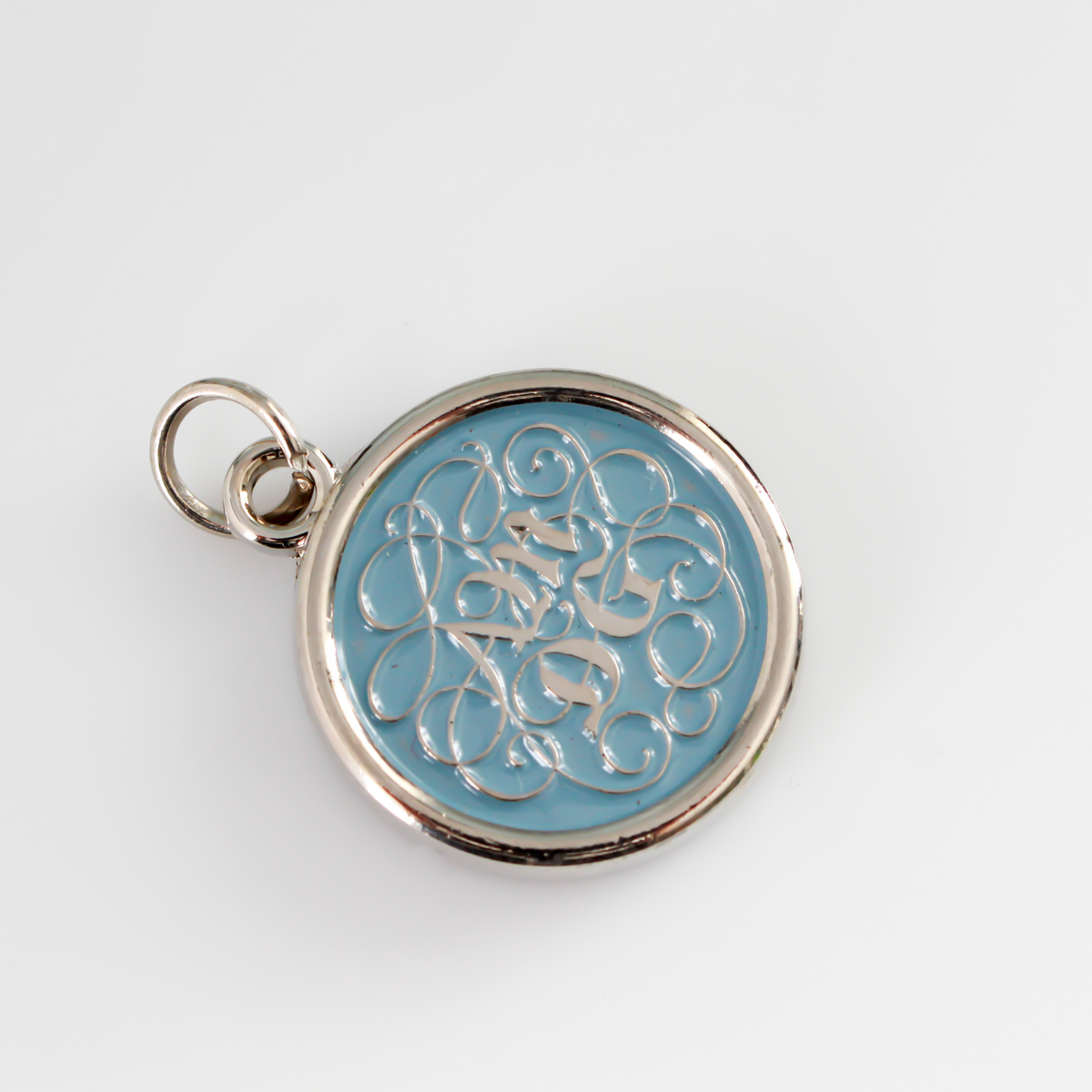 Round pendant with an ornate acronym on the front that is detailed by a light blue enamel color. The acronym ADMG stands for "Ad Majorem Dei Gloriam".