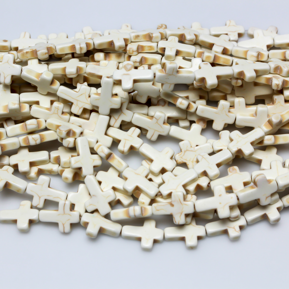 Beige cross shaped beads that are a synthetic turquoise material dyed an off-white beige color. Sold by the strand