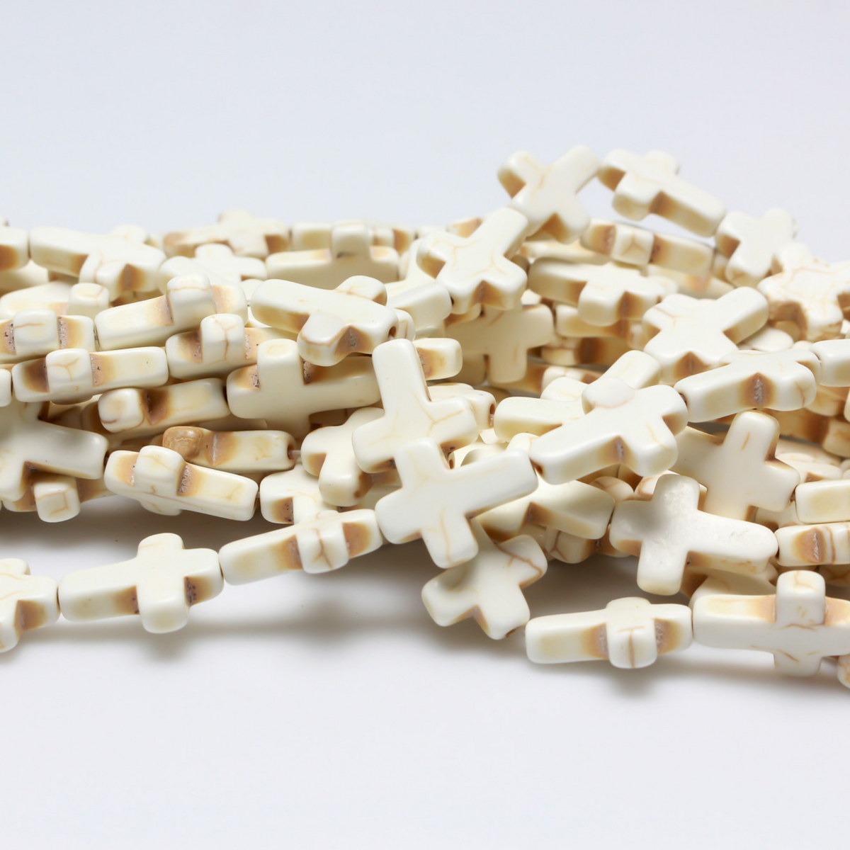 Beige cross shaped beads that are a synthetic turquoise material dyed an off-white beige color. Sold by the strand