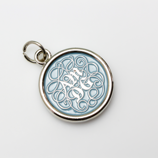Round pendant with an ornate acronym on the front that is detailed by a light blue enamel color. The acronym ADMG stands for "Ad Majorem Dei Gloriam".