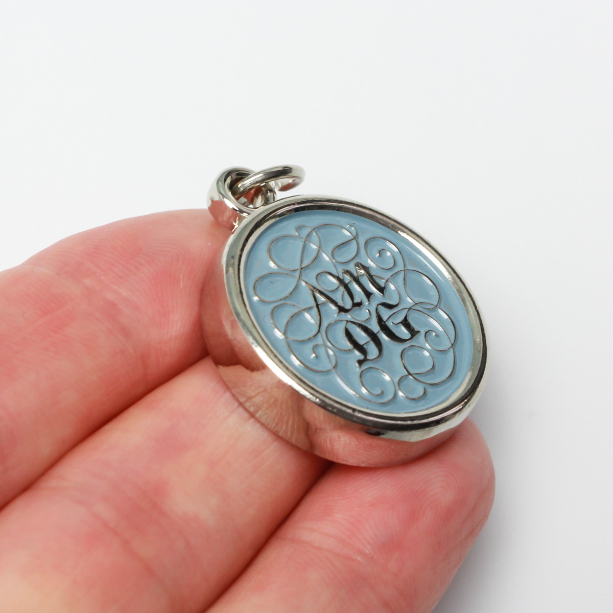 Round pendant with an ornate acronym on the front that is detailed by a light blue enamel color. The acronym ADMG stands for "Ad Majorem Dei Gloriam".