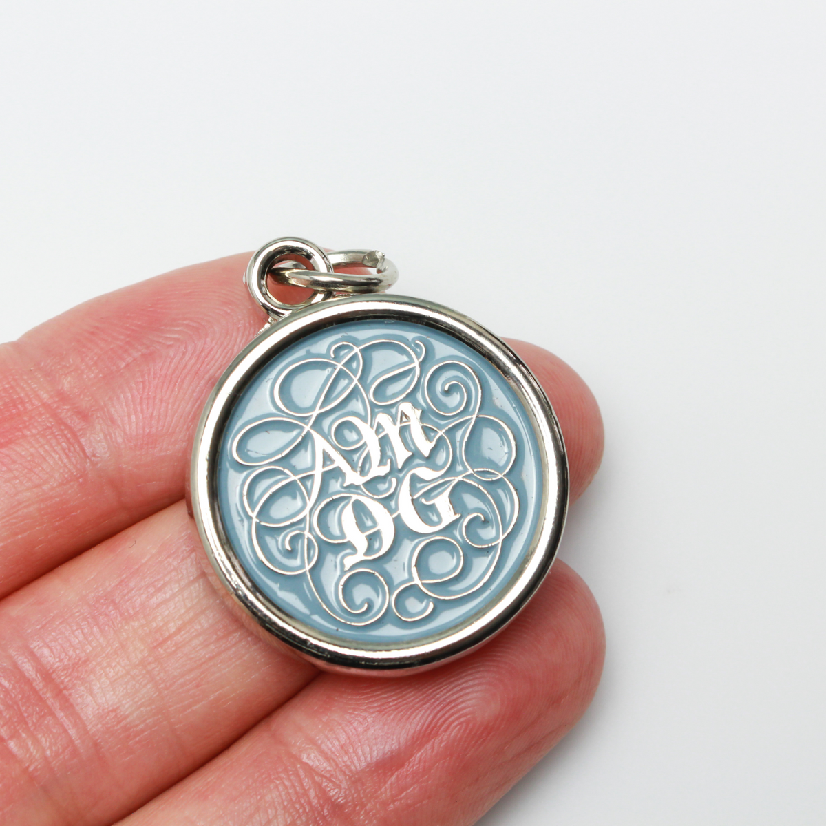 Round pendant with an ornate acronym on the front that is detailed by a light blue enamel color. The acronym ADMG stands for "Ad Majorem Dei Gloriam".