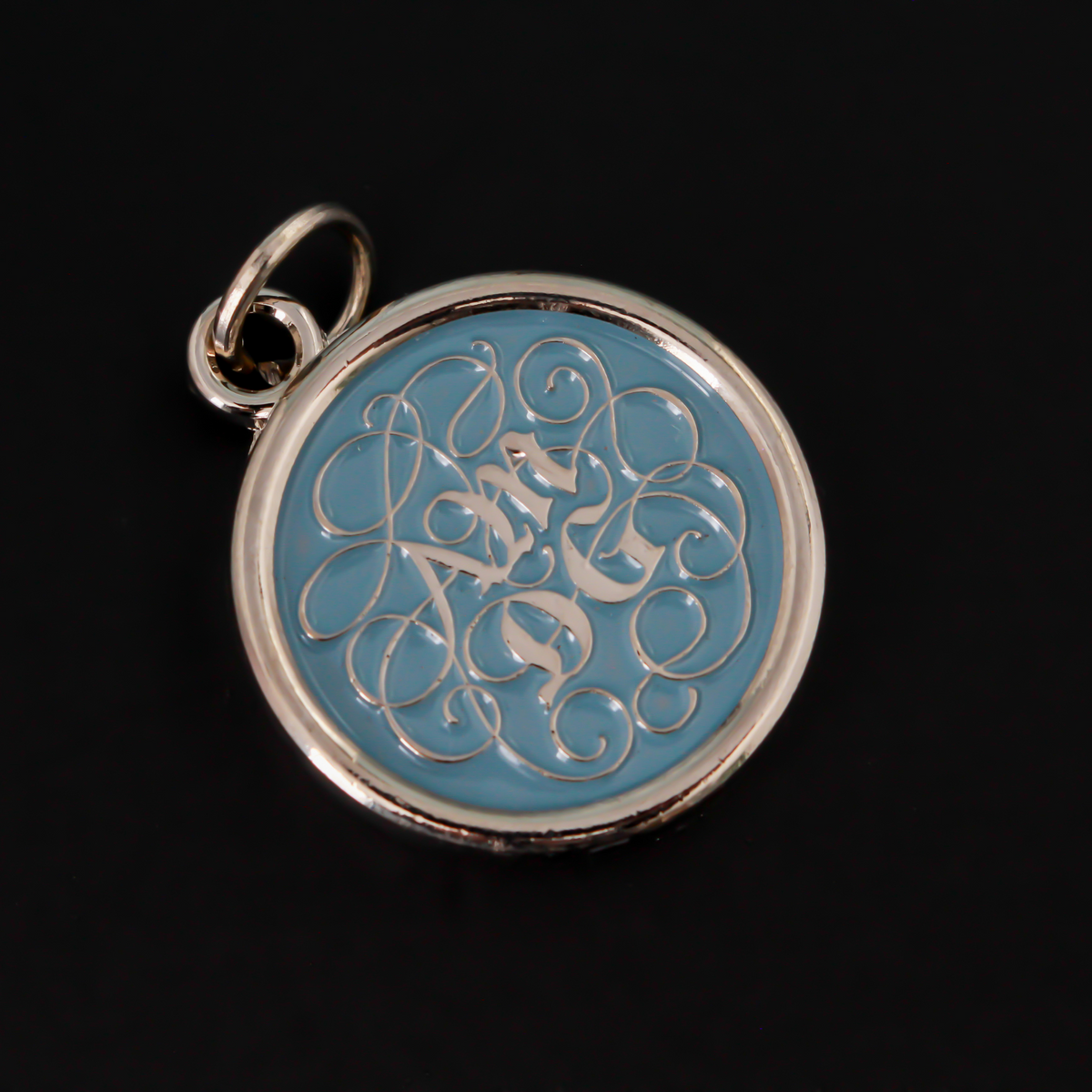 Round pendant with an ornate acronym on the front that is detailed by a light blue enamel color. The acronym ADMG stands for "Ad Majorem Dei Gloriam".