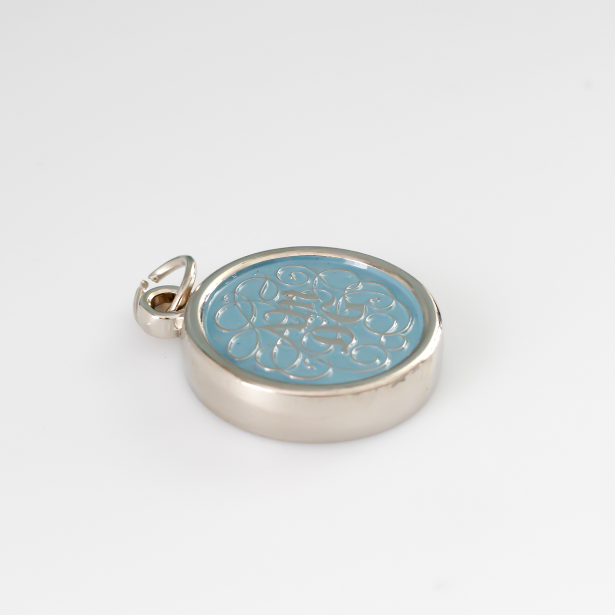 Round pendant with an ornate acronym on the front that is detailed by a light blue enamel color. The acronym ADMG stands for "Ad Majorem Dei Gloriam".