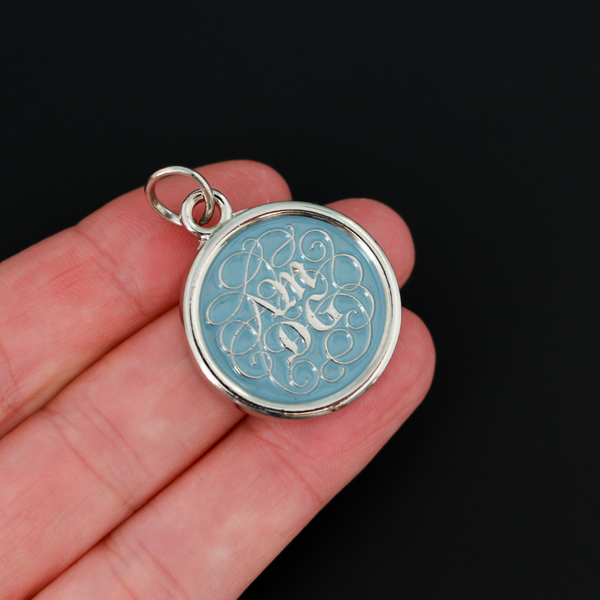 Round pendant with an ornate acronym on the front that is detailed by a light blue enamel color. The acronym ADMG stands for "Ad Majorem Dei Gloriam".