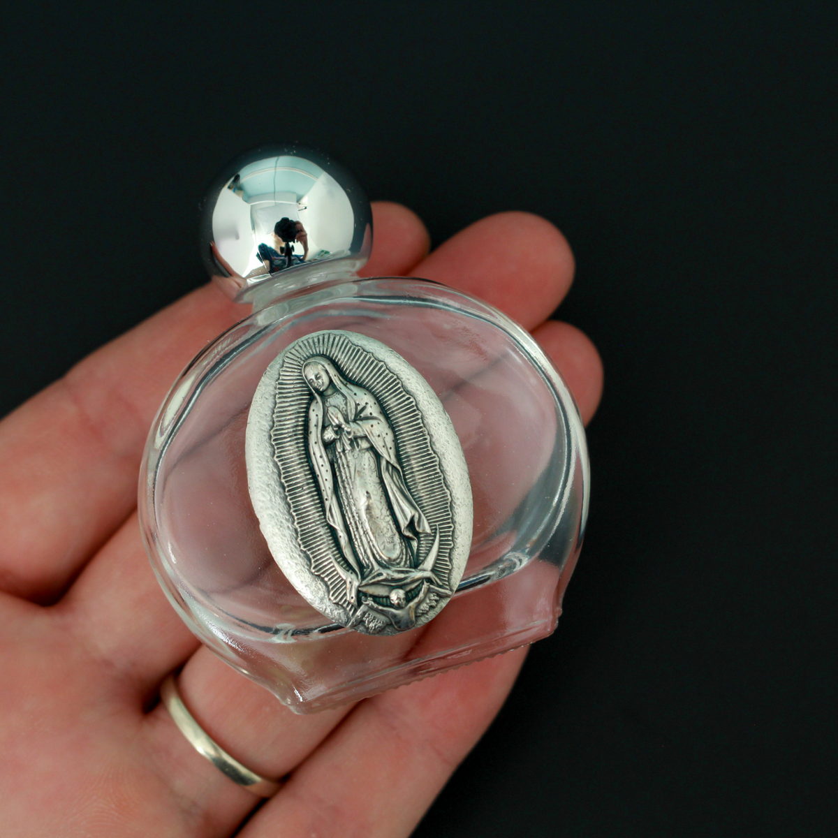 Glass holy water bottle with a metal embossed plaque of Our Lady of Guadalupe on the front. Plastic screw on cap that is a shiny silver tone color