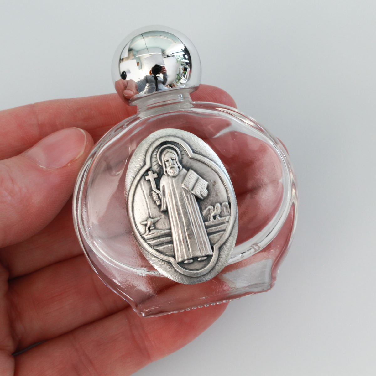 Glass holy water bottle with a metal embossed plaque of Saint Benedict on the front. Plastic screw on cap that is a shiny silver tone color.