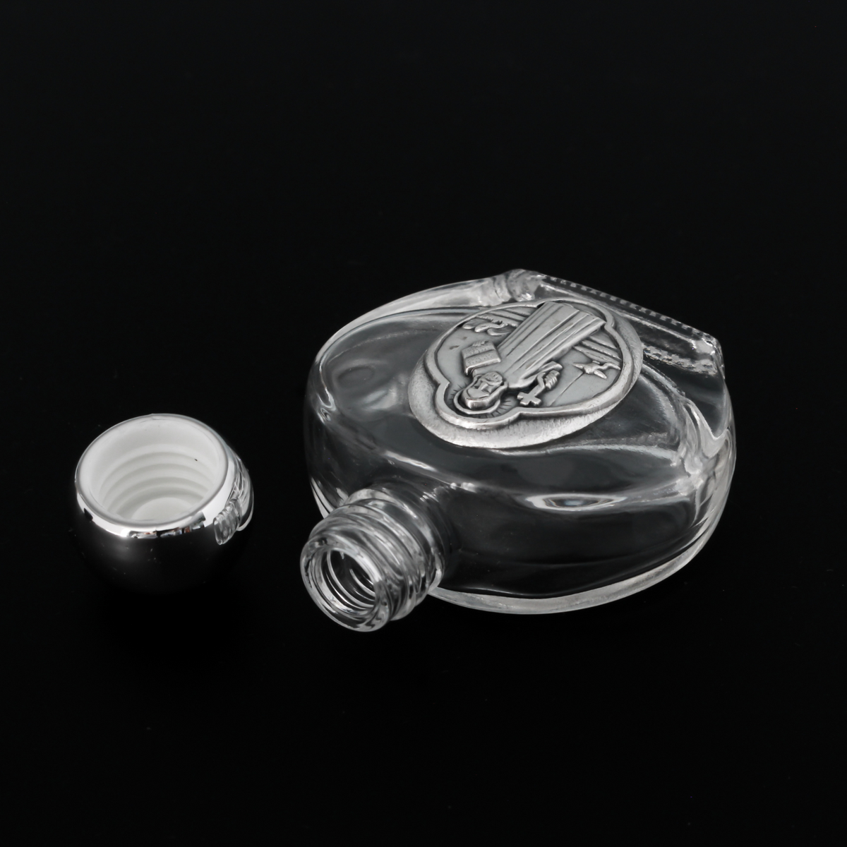 Glass holy water bottle with a metal embossed plaque of Saint Benedict on the front. Plastic screw on cap that is a shiny silver tone color.