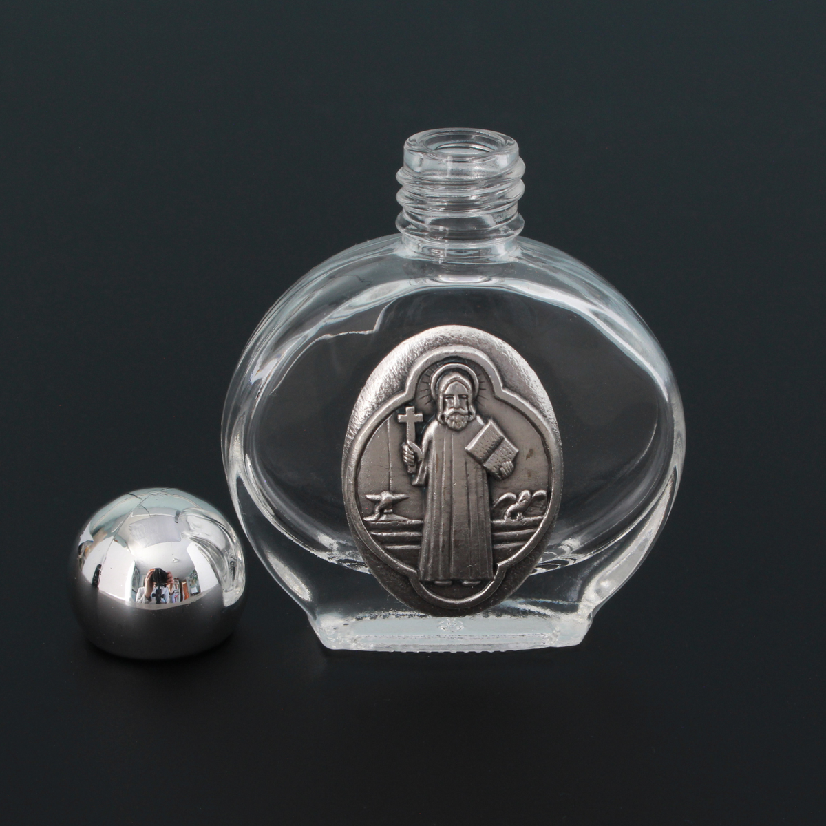 Glass holy water bottle with a metal embossed plaque of Saint Benedict on the front. Plastic screw on cap that is a shiny silver tone color.