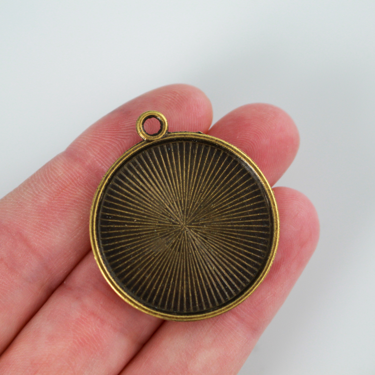 Round pendant cabochon setting in an antiqued bronze color. There is a pattern along the edge of the cup. The tray fits 25mm cabochons