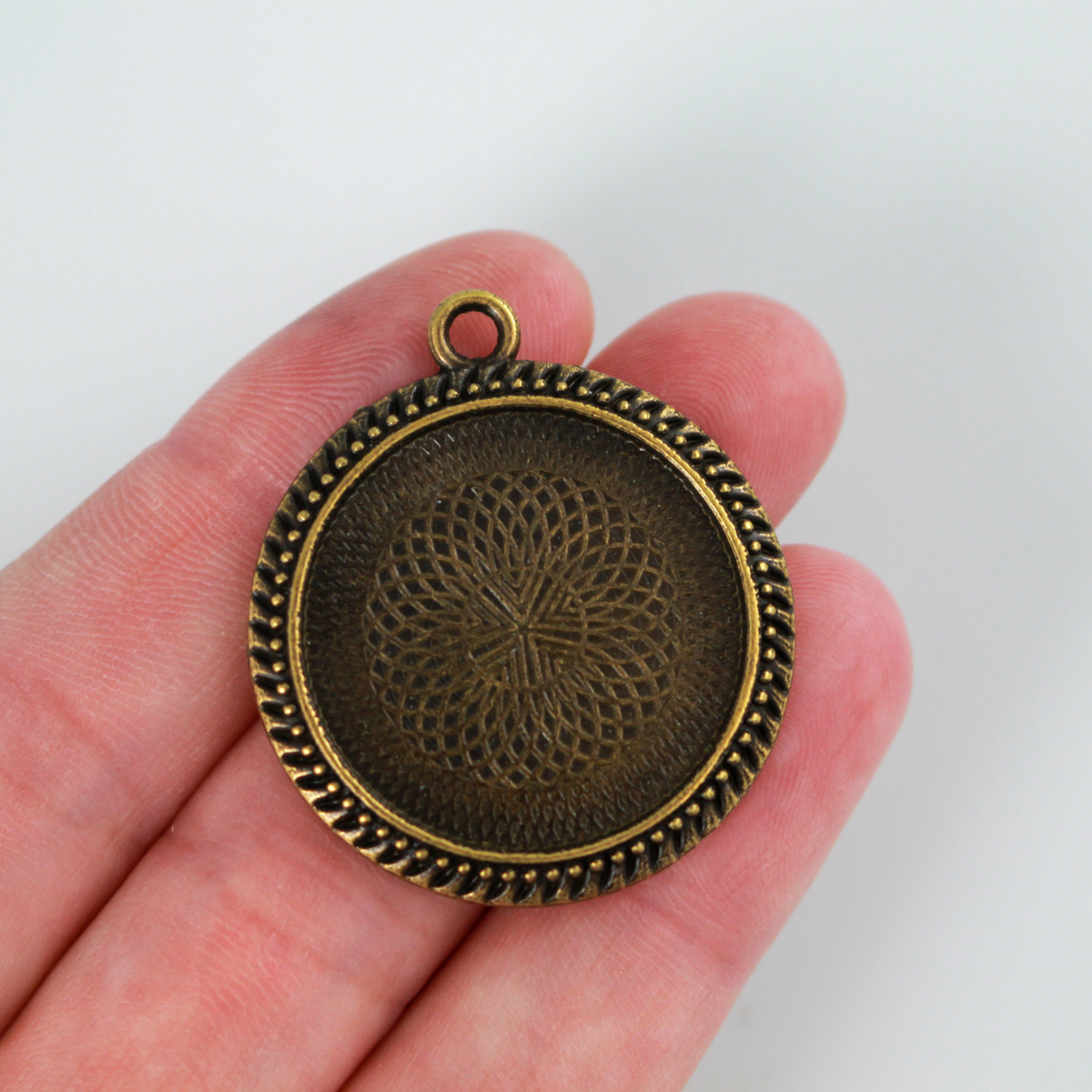 Round pendant cabochon setting in an antiqued bronze color. There is a pattern along the edge of the cup. The tray fits 25mm cabochons