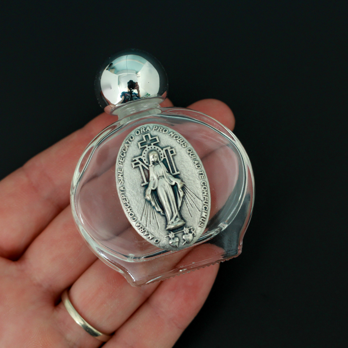 Glass holy water bottle with a metal embossed plaque of The Miraculous Medal on the front. Plastic screw on cap that is a shiny silver tone color.